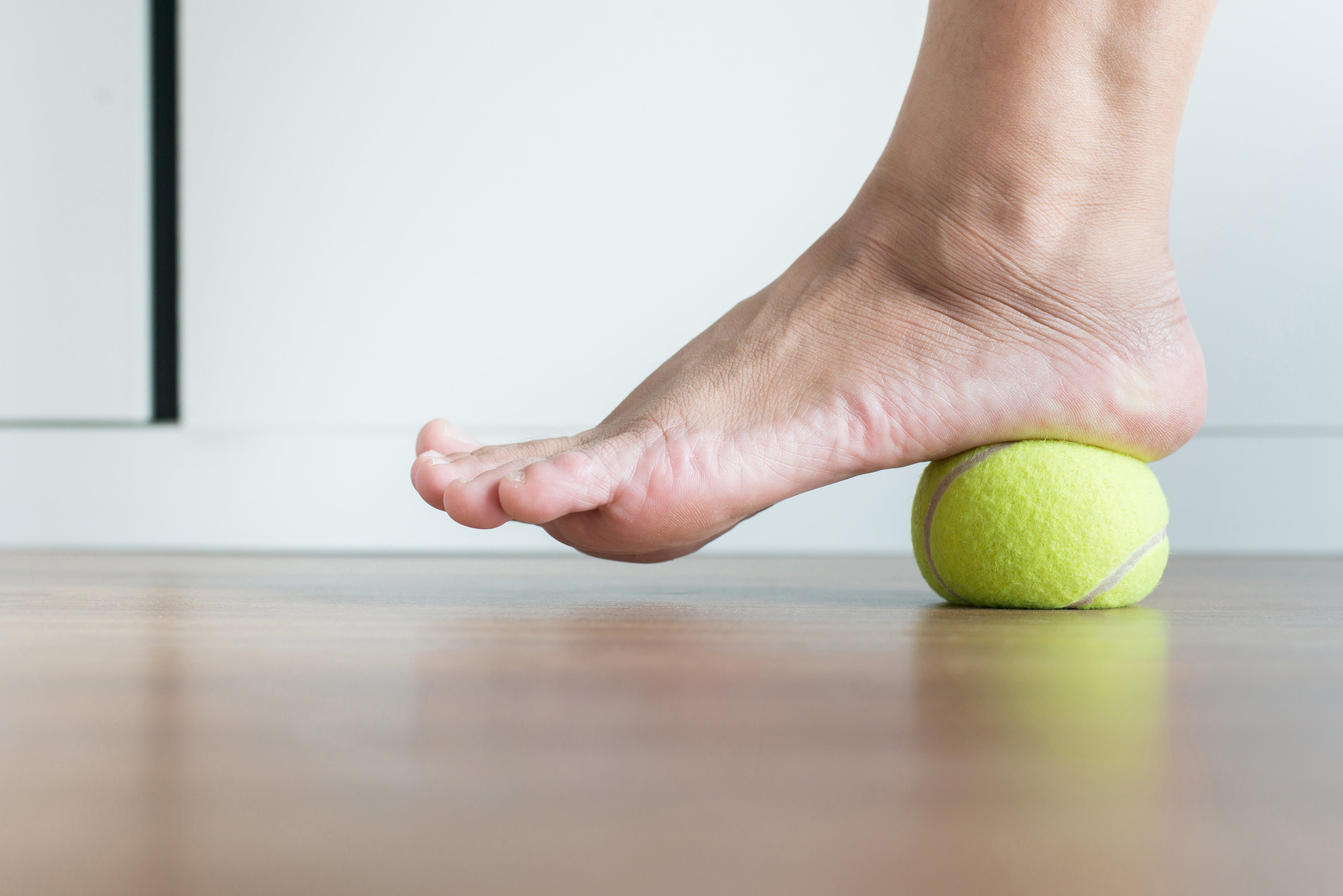 Person performing a plantar fascia massage by rolling a tennis ball under the arch of the foot to relieve heel pain and tension.