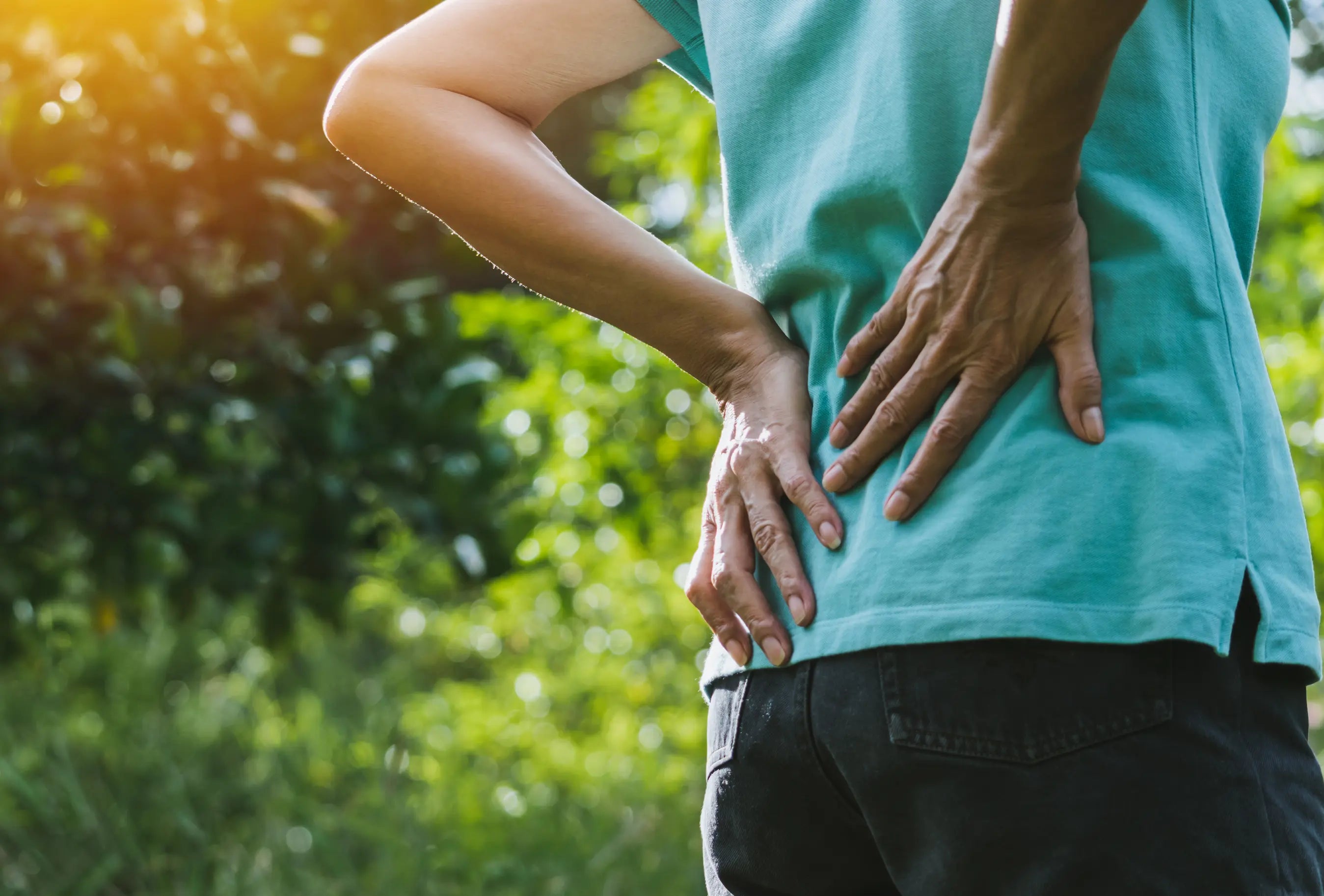 Person outdoors holding their lower back with both hands, showing back pain or discomfort while standing in a green, sunlit park setting.