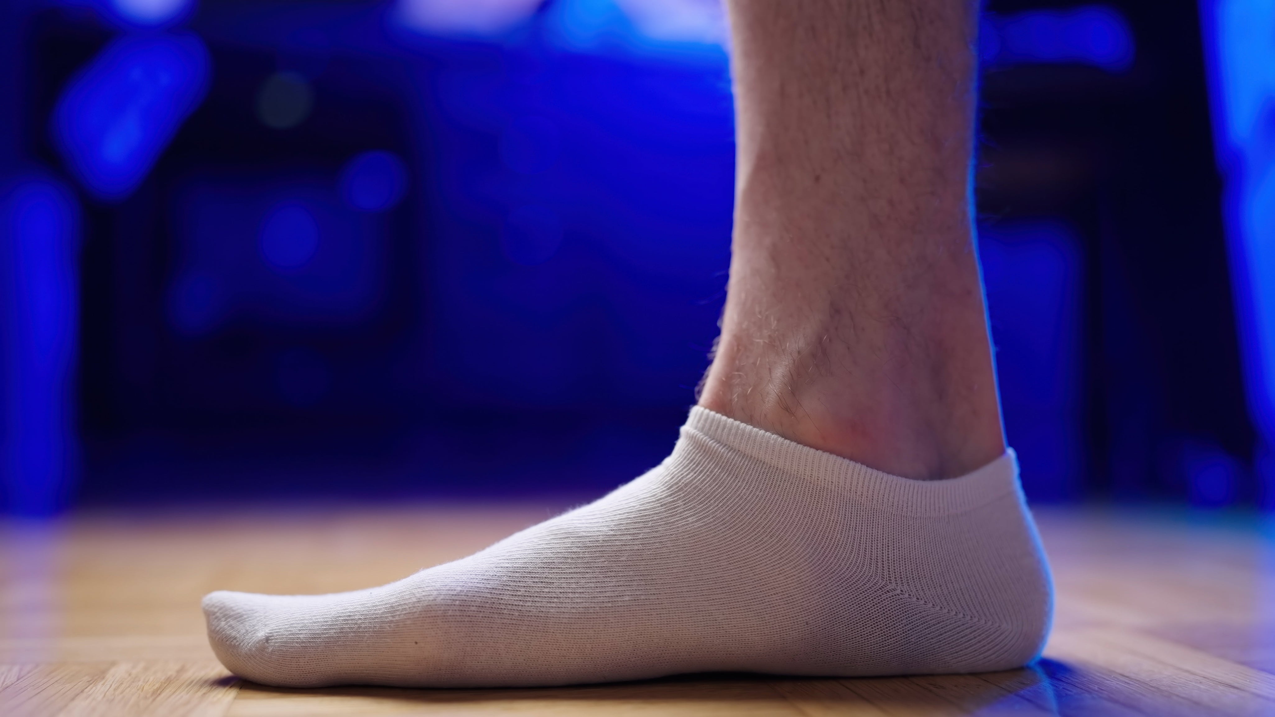 A close-up side view of a person’s foot in a low-cut white sock, standing on a hardwood floor with a visible low arch, against a blurred blue background.