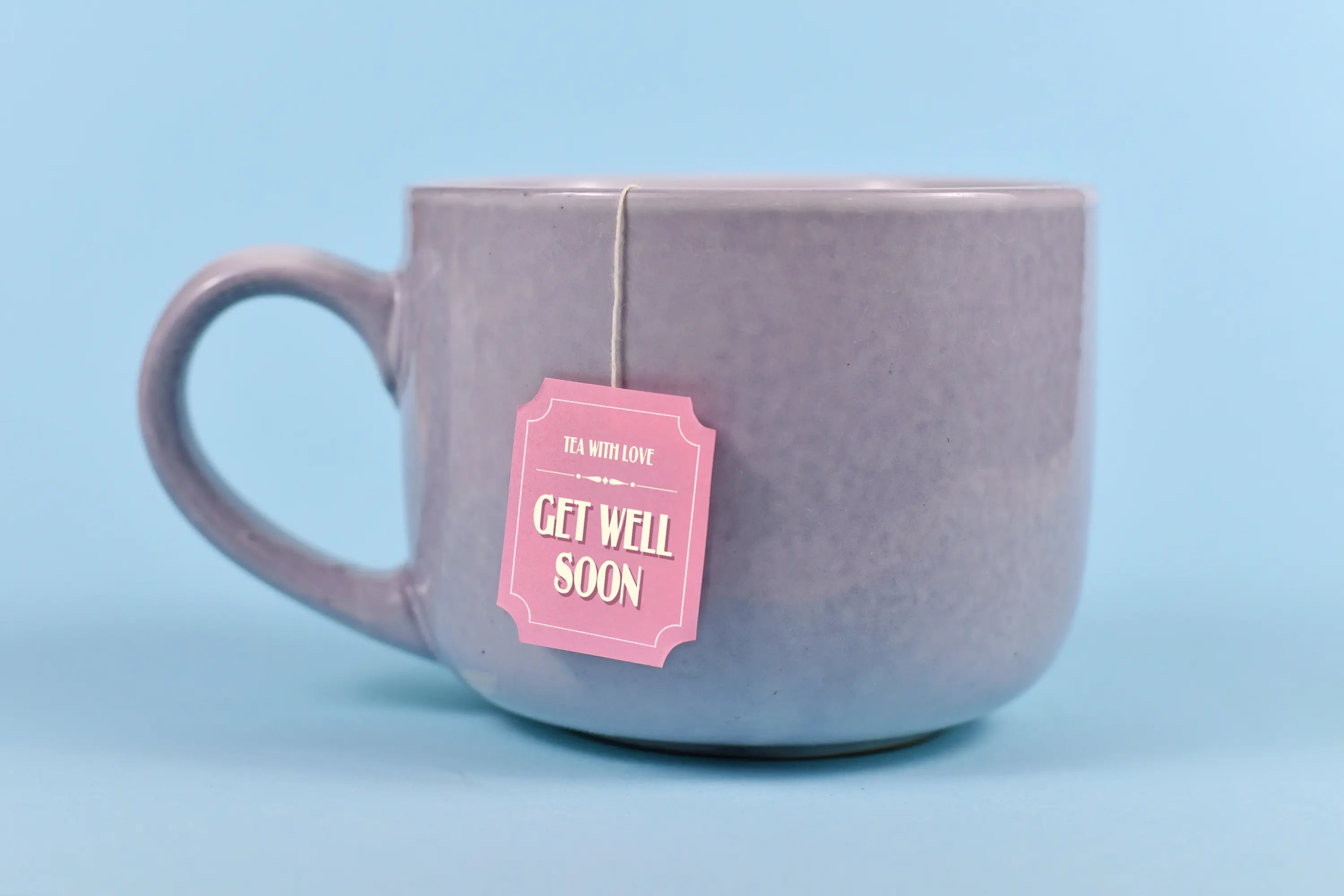 Gray ceramic mug with a tea bag tag that reads “Get Well Soon” against a soft blue background.