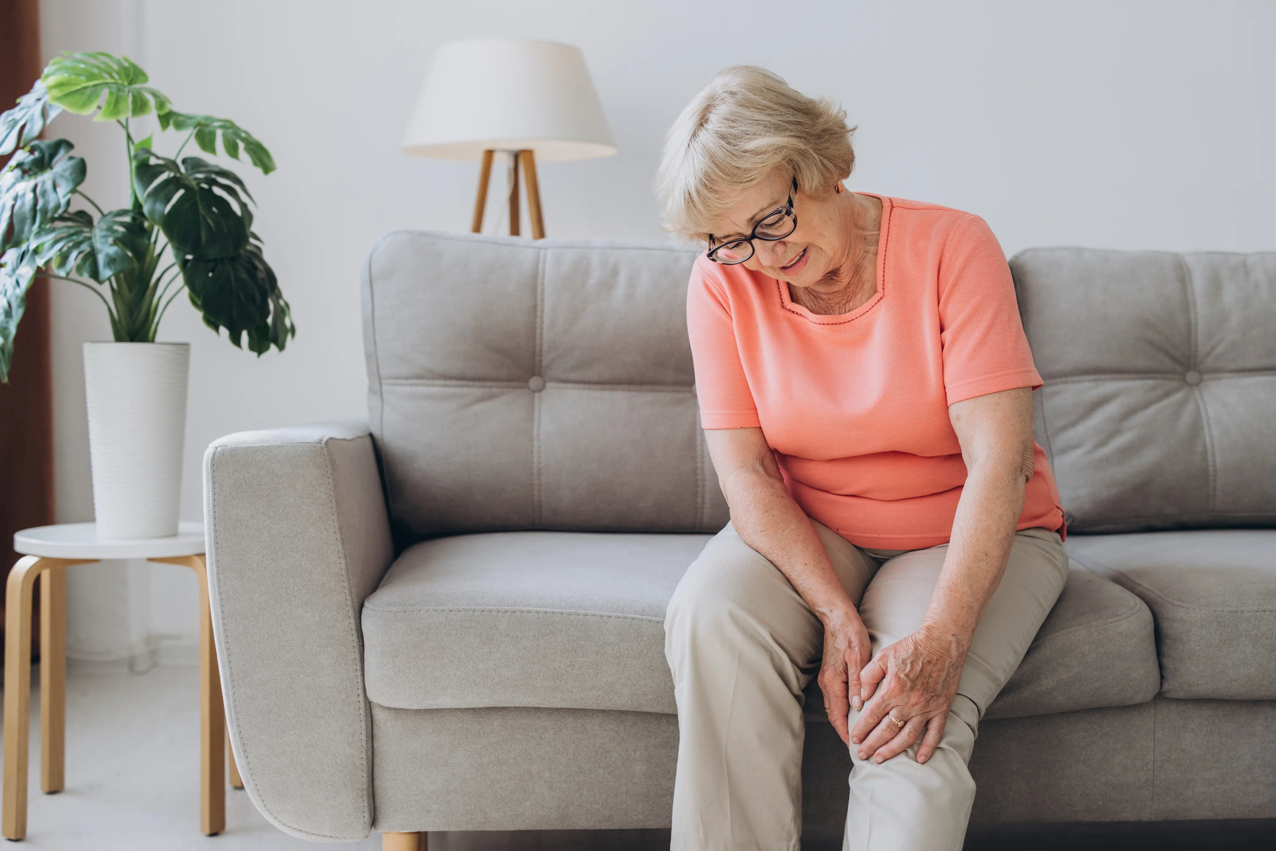Older woman sitting on a couch holding her knee with a pained expression, experiencing knee discomfort in a home living room setting.