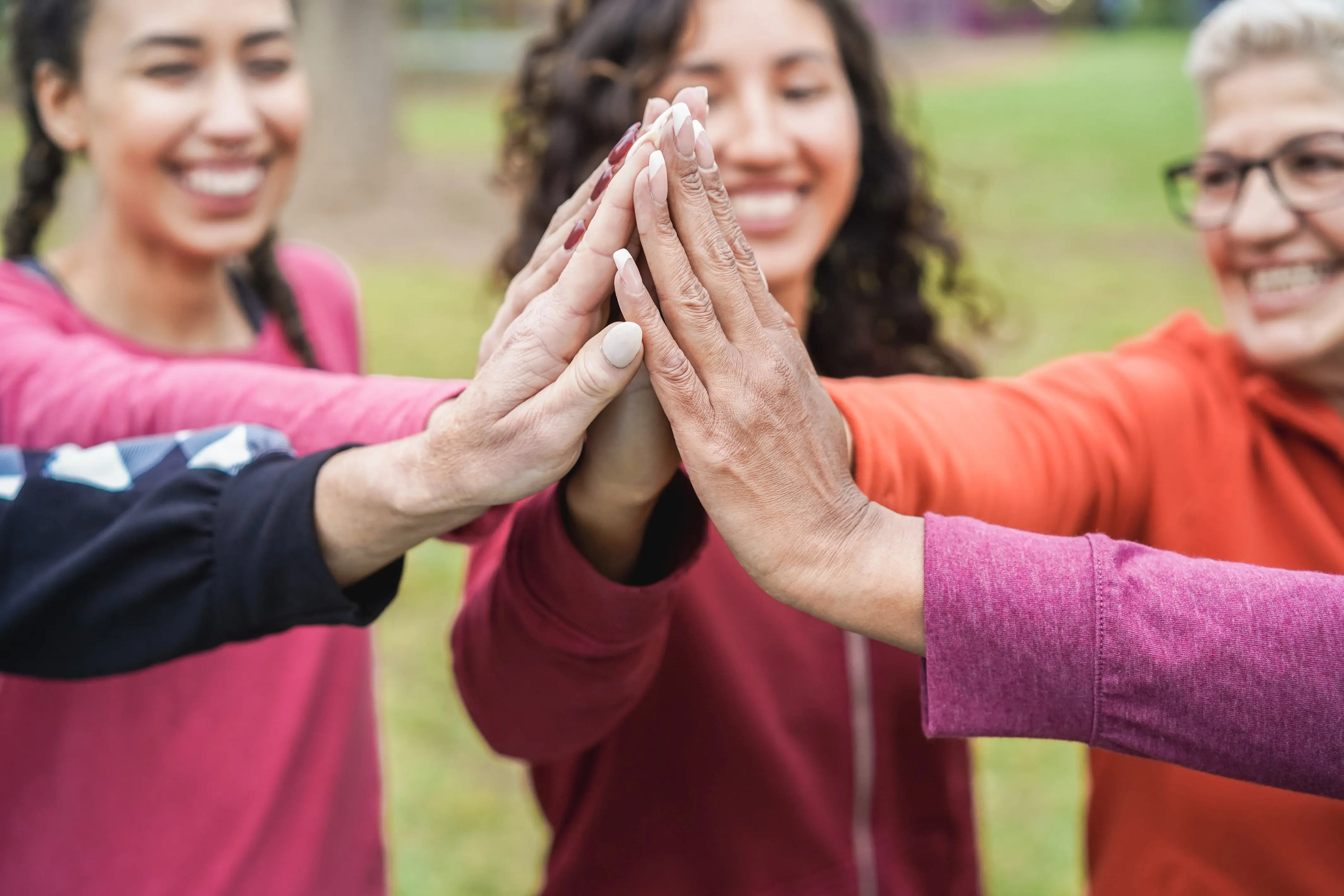 A group of diverse women smiling and stacking their hands together in a supportive high-five gesture outdoors, symbolizing community and encouragement.