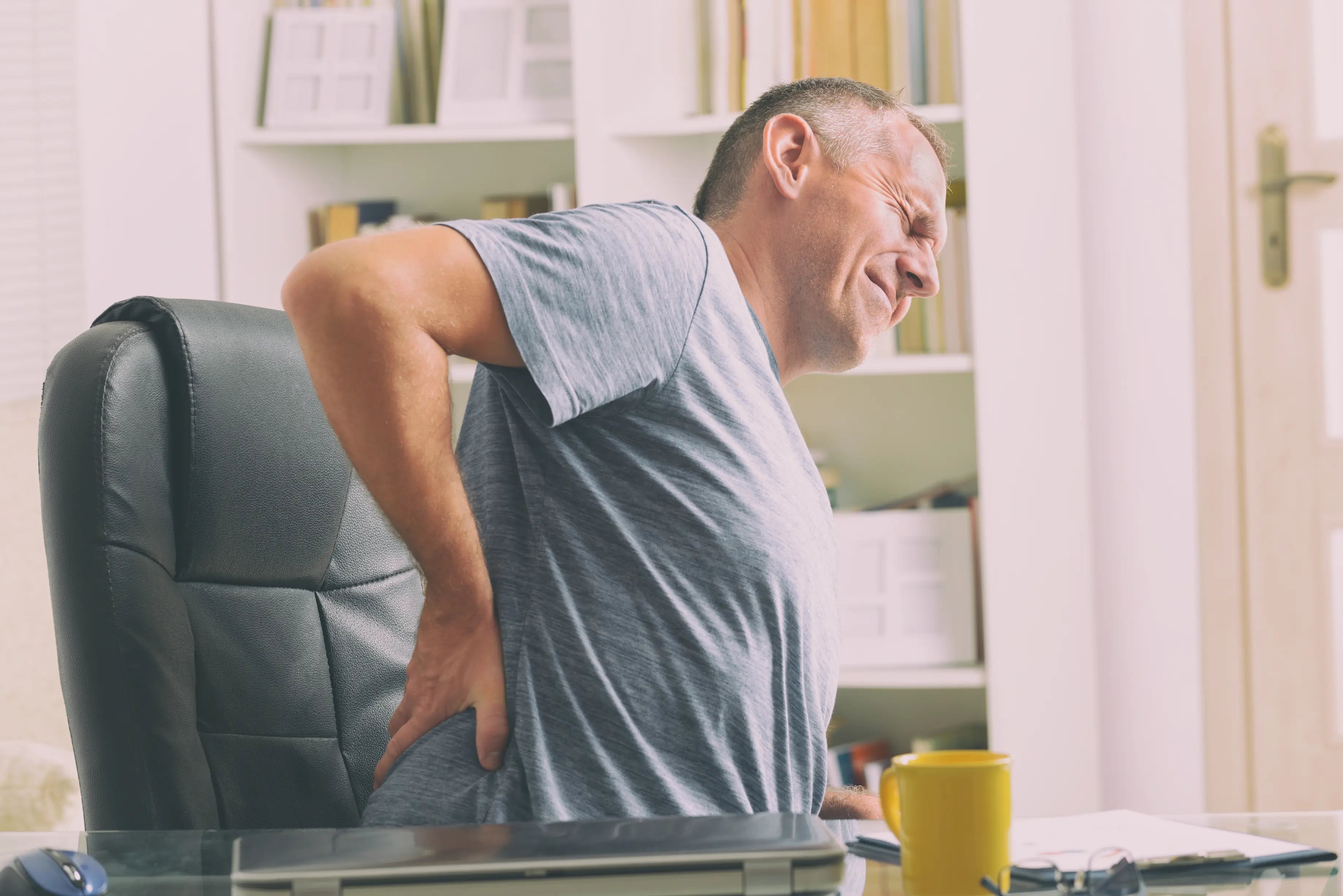 Middle-aged man sitting in an office chair and holding his lower back in pain, showing discomfort from back pain while working at a desk.
