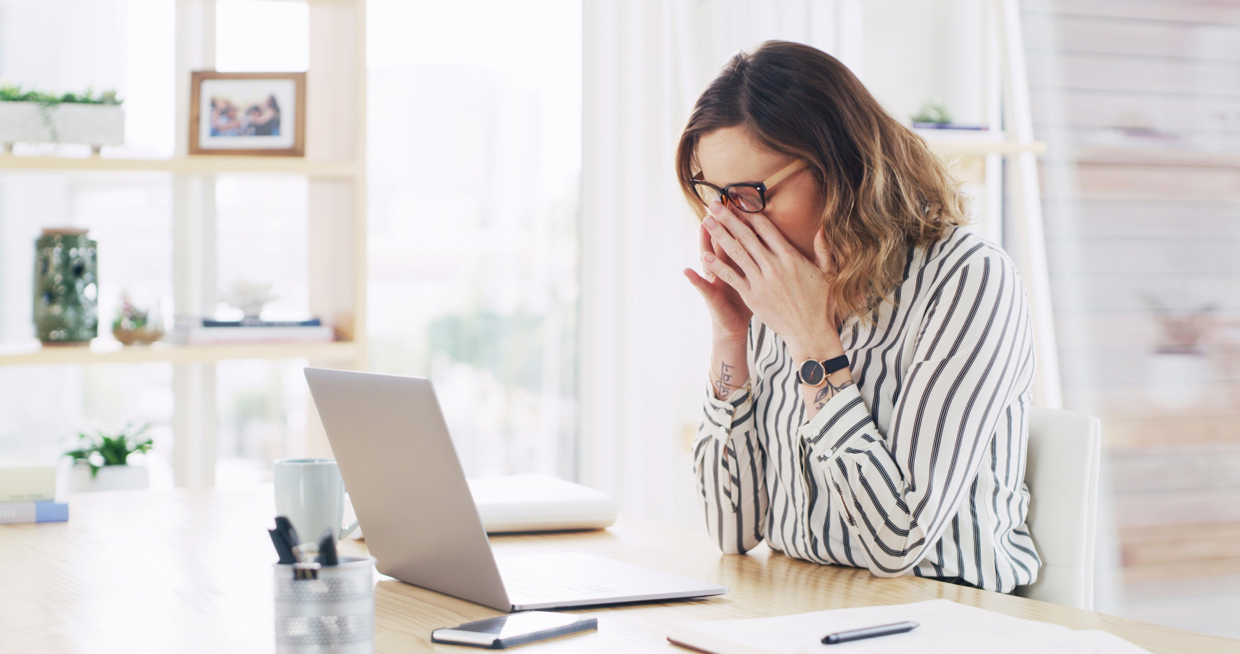 Woman experiencing menopause-related brain fog, sitting at a desk with her hands on her face while working on a laptop, appearing tired and overwhelmed.