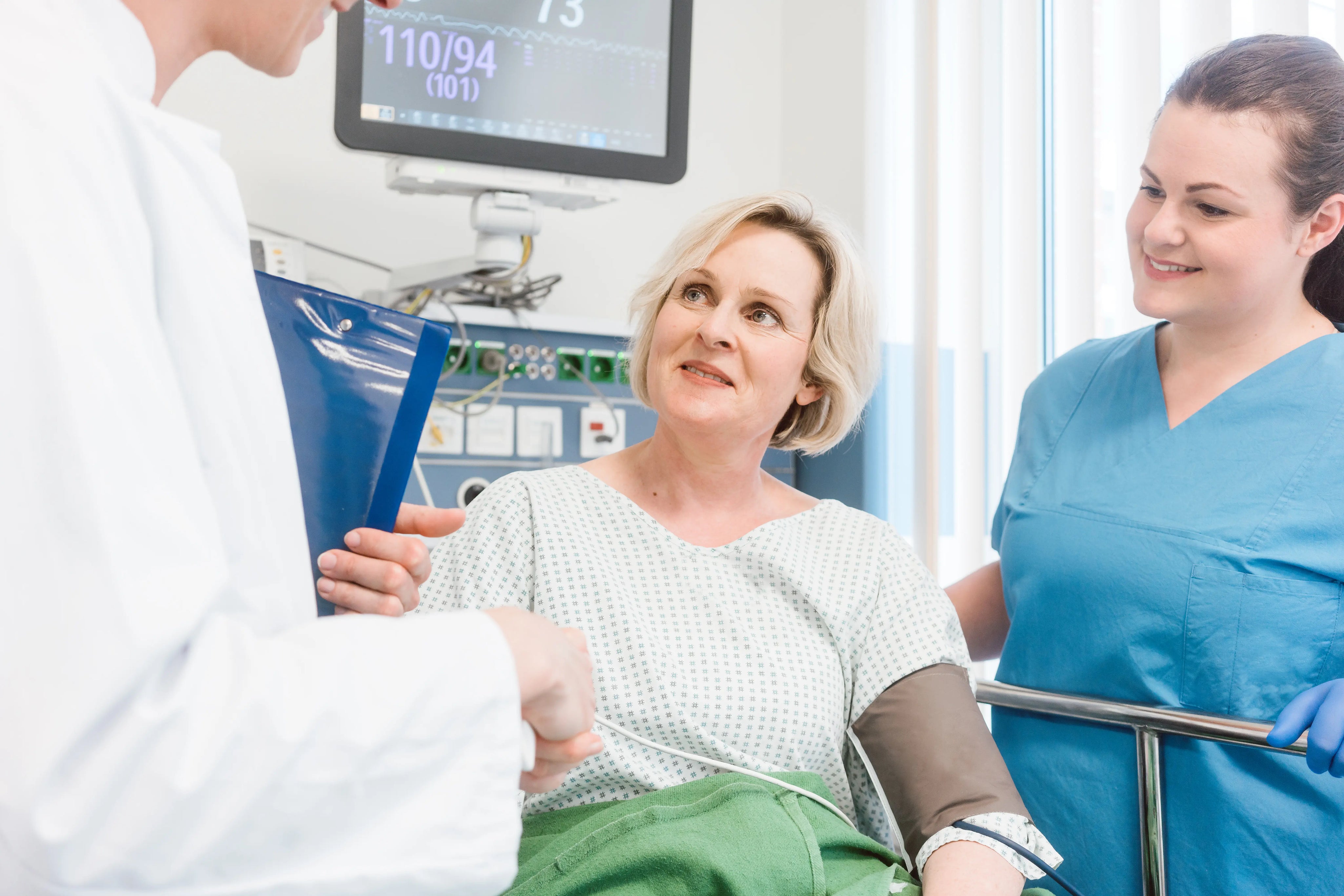 Female patient in hospital bed speaking with doctor and nurse during post-surgery recovery checkup with medical monitor in background