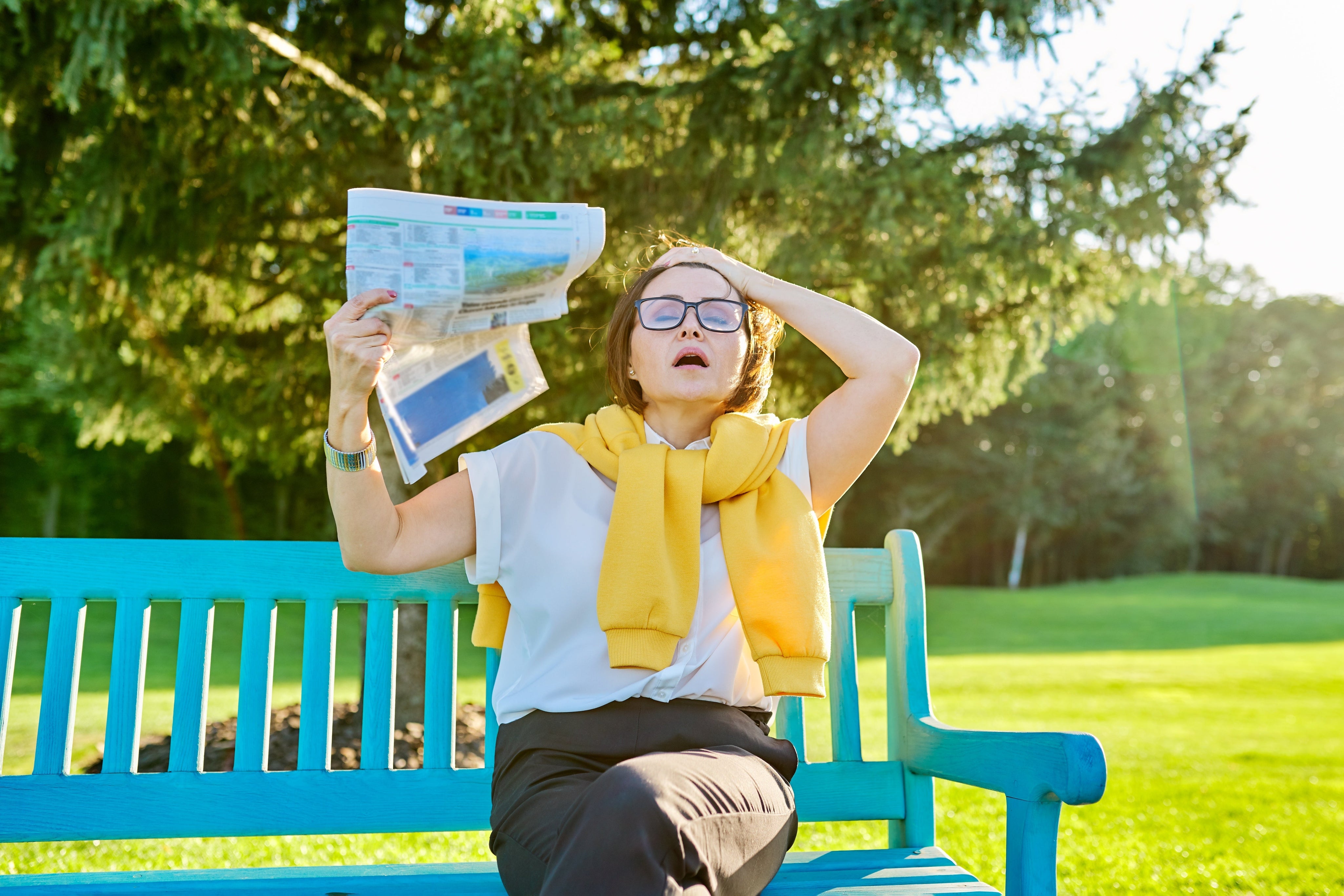 A middle-aged woman sitting on a bench outdoors, fanning herself with a newspaper and looking overheated, illustrating a hot flash during perimenopause.