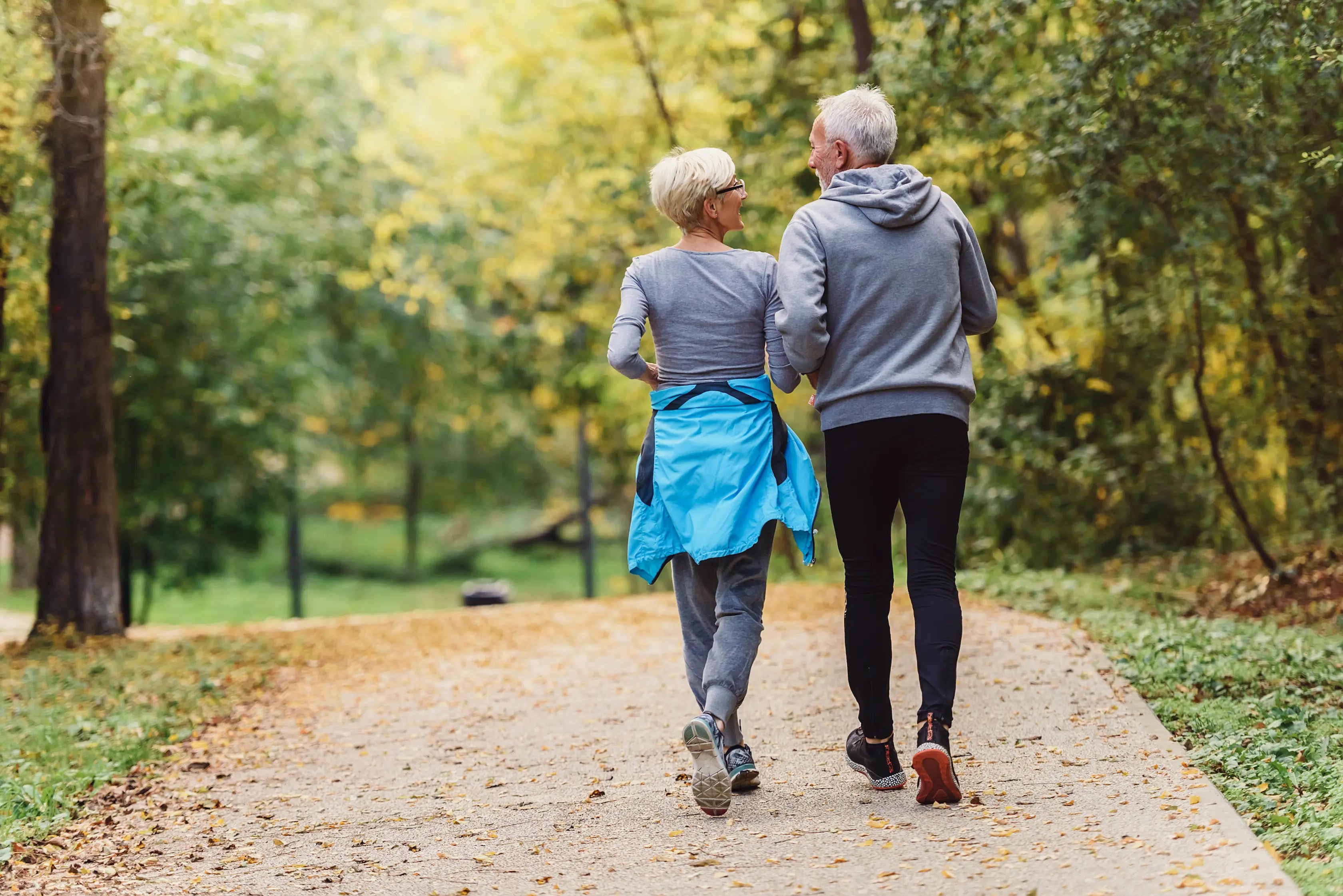 An older couple walking arm-in-arm along a paved park path surrounded by lush green and yellow autumn foliage