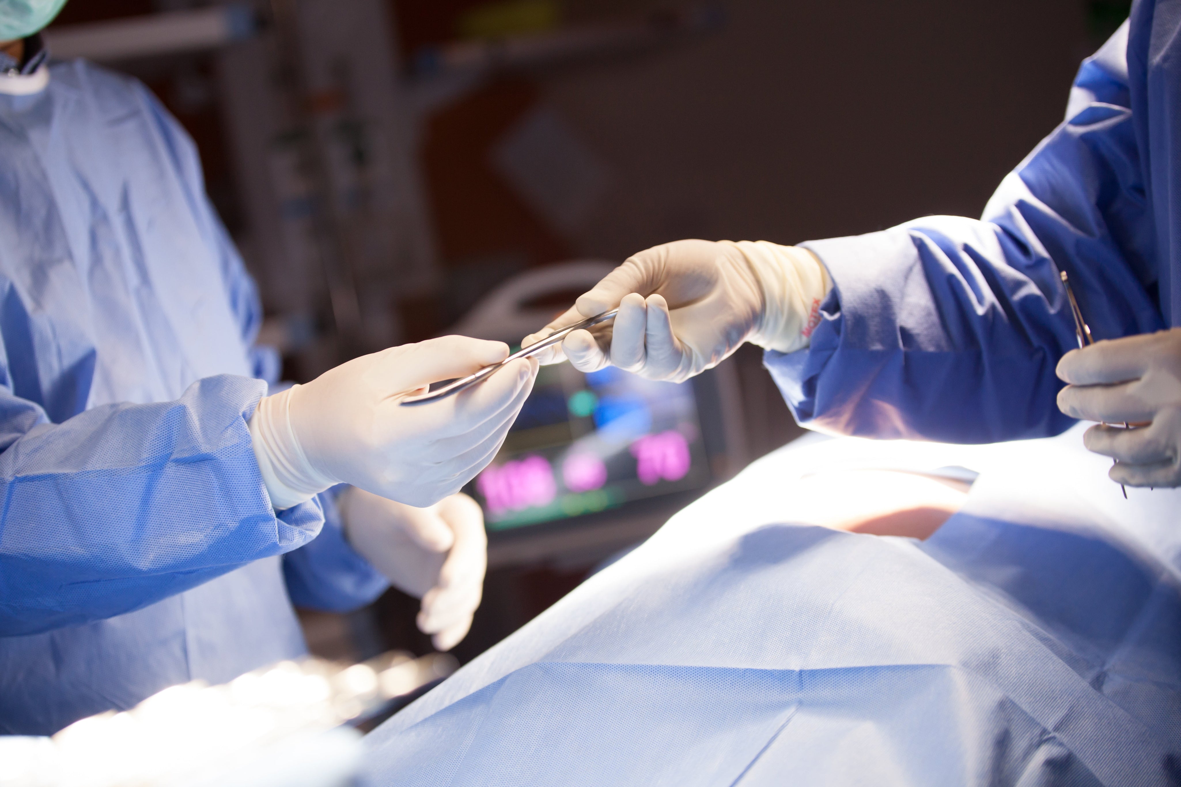 Two surgeons passing a surgical instrument during an operation in a hospital operating room with patient draped and monitors in background