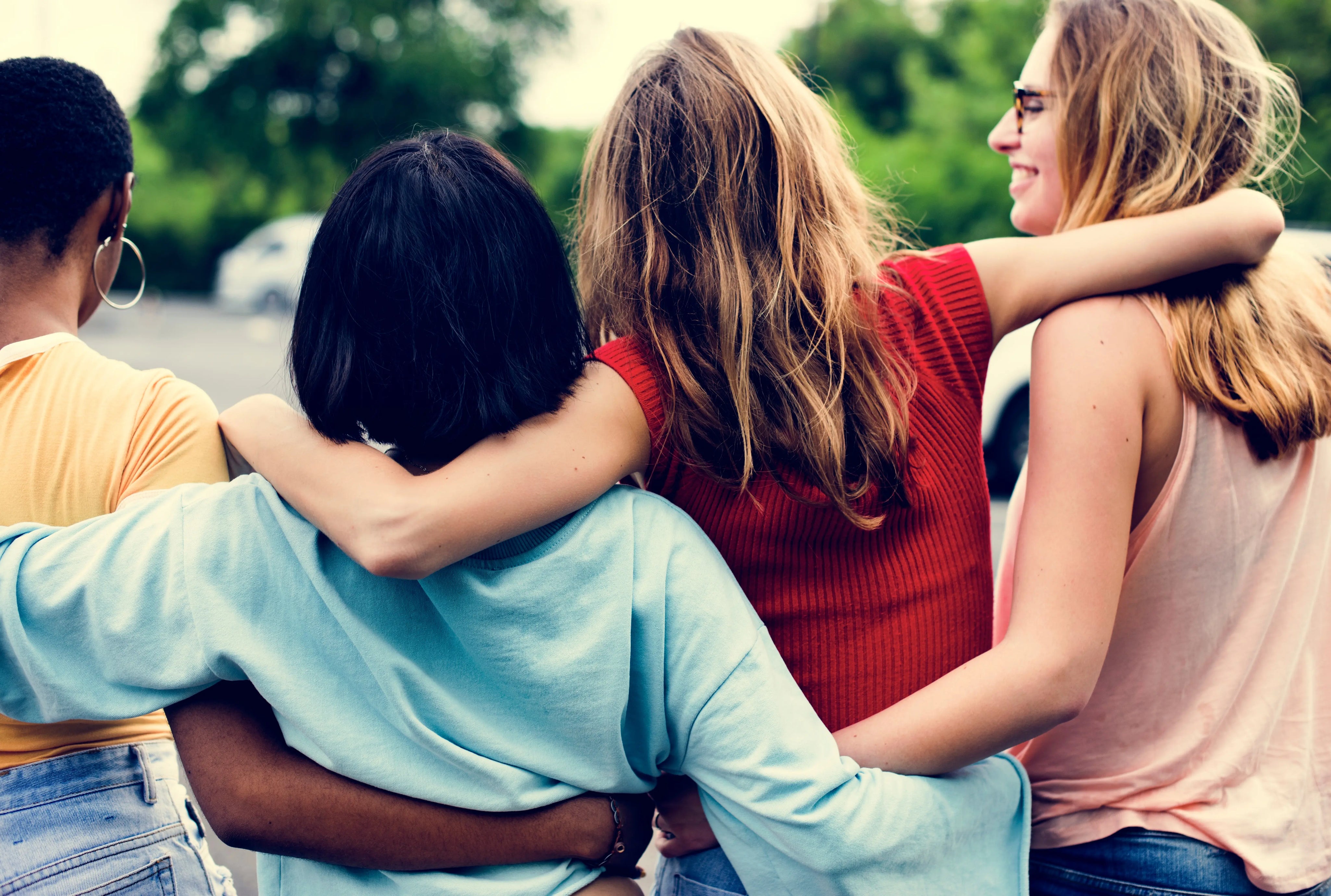 Group of women standing outdoors with their arms around each other, viewed from behind, showing support and friendship.