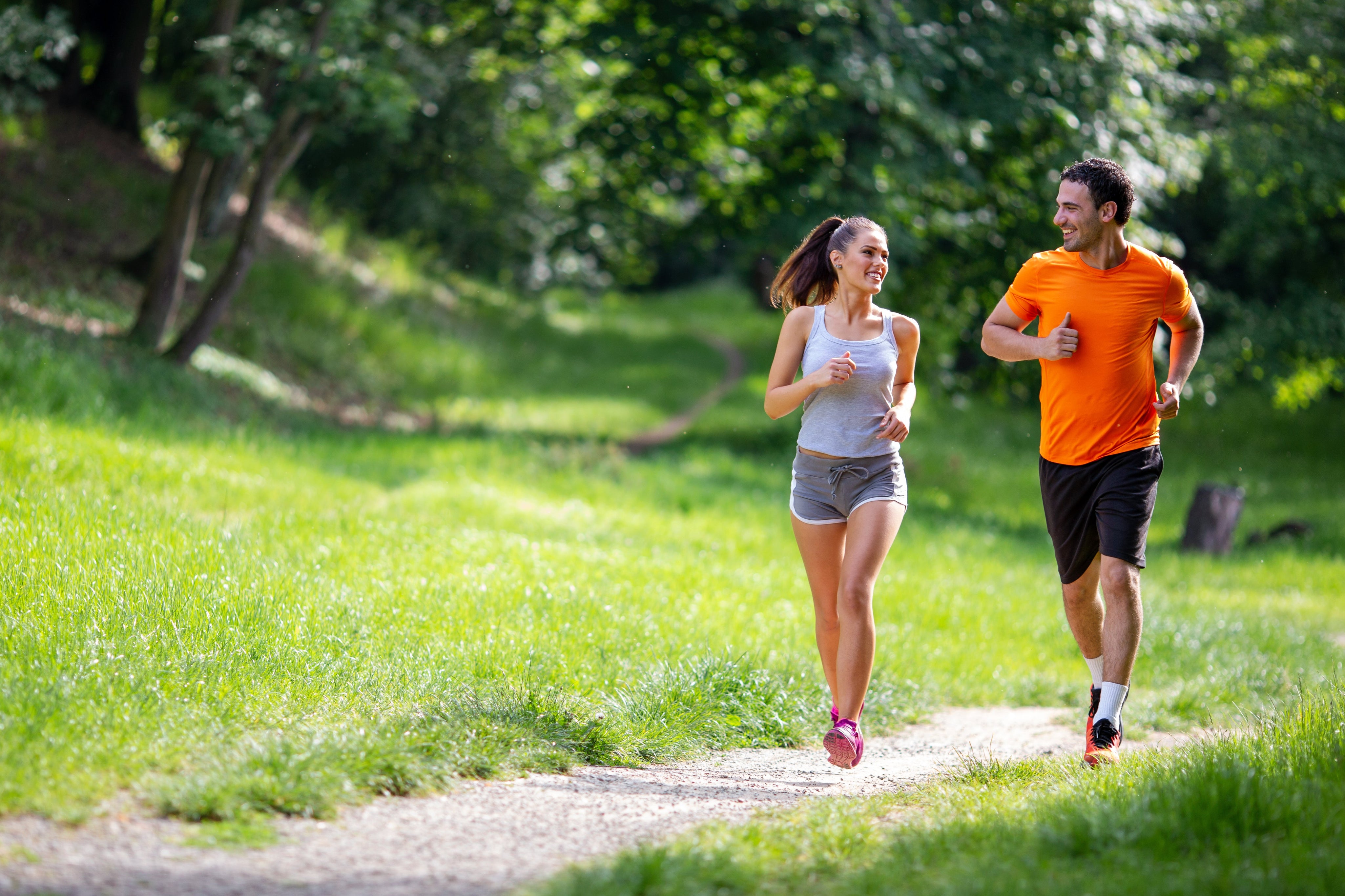 A smiling young man and woman jogging together along a sunny park trail surrounded by lush green grass and trees