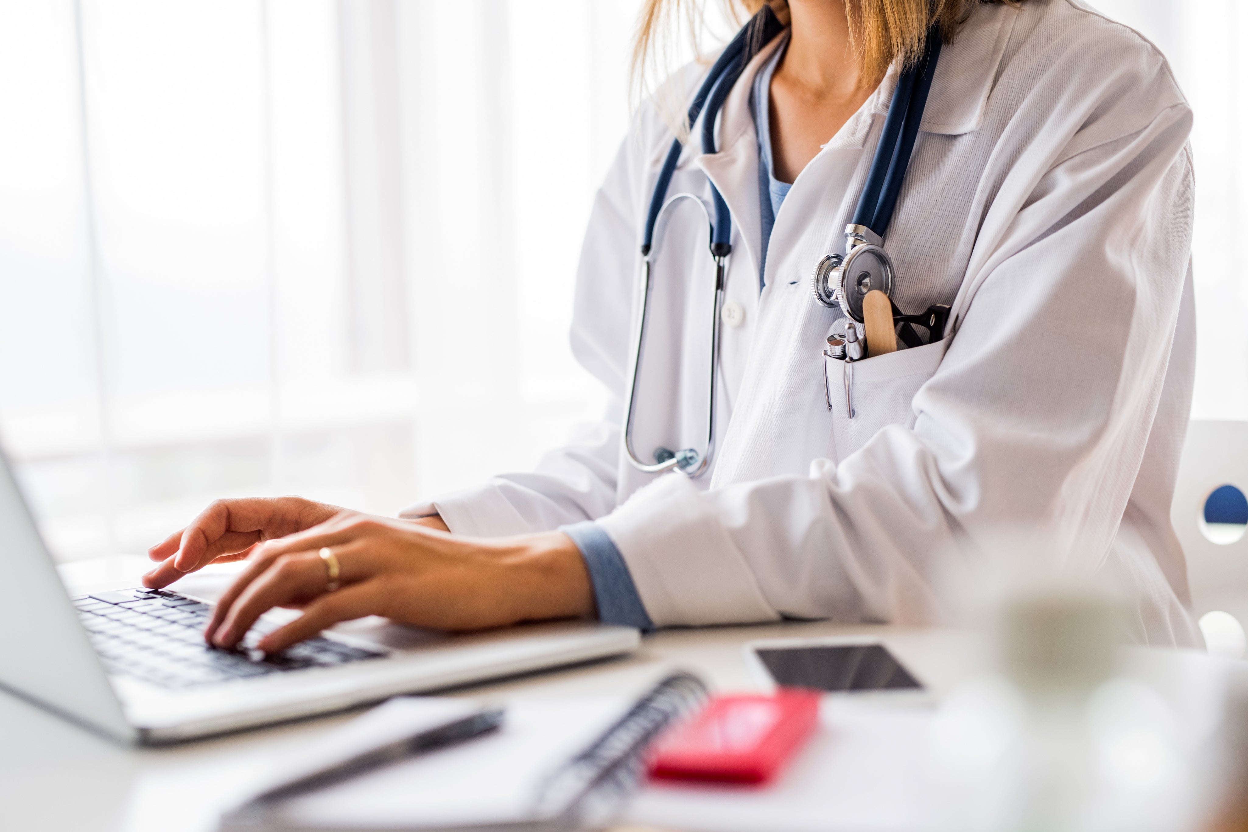 Healthcare provider in a white coat typing on a laptop at a desk, wearing a stethoscope and reviewing patient information in a clinical workspace.